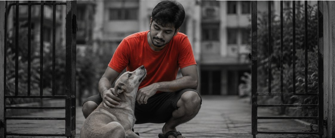 A young man crouched at an open gate, one hand on the head of a street dog looking up at him.