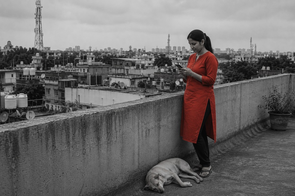 A woman in a red kurta reading her phone on a rooftop terrace in flat daylight, a street dog asleep at her feet.